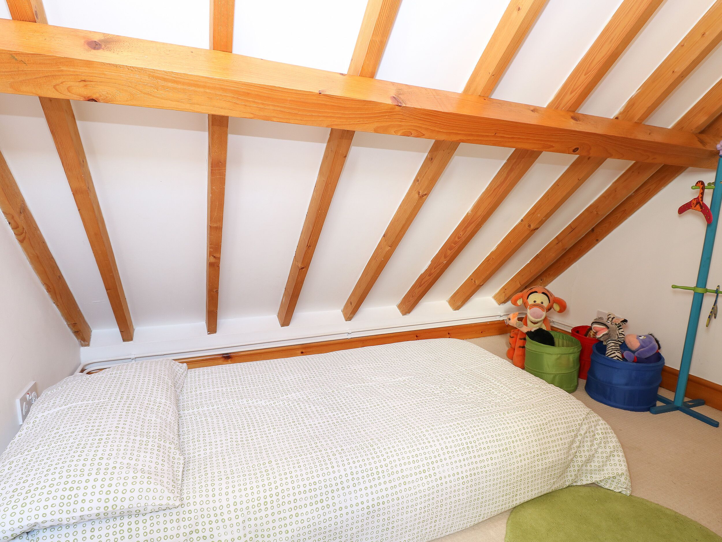 Una stanza con soffitto a falde sostenuto da travi in legno, un letto con copriletto decorato e diversi peluche e contenitori portaoggetti.