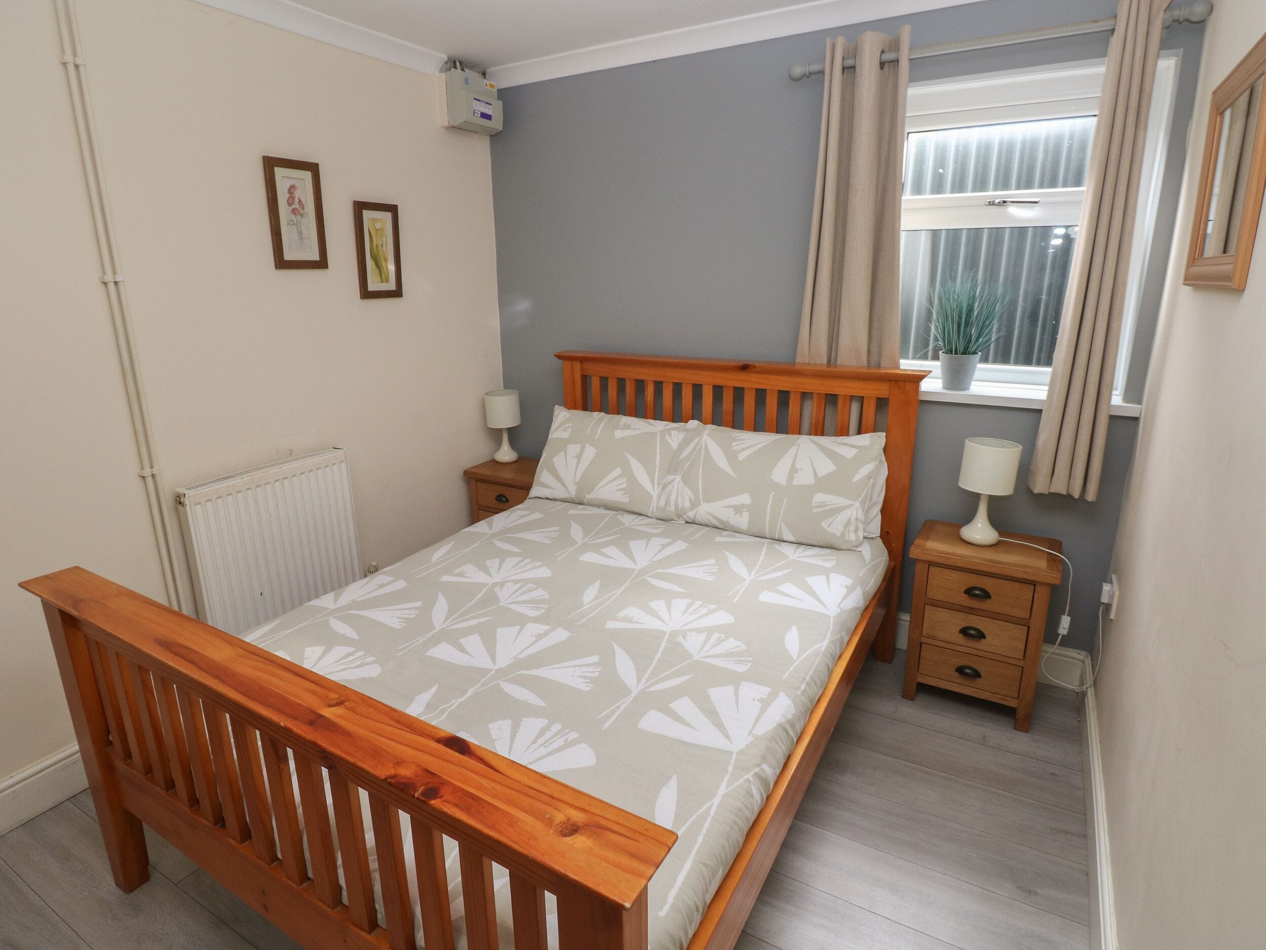 A bedroom with a wooden bed, two bedside tables with lamps, a radiator, a window with curtains, and two framed pictures on the wall.