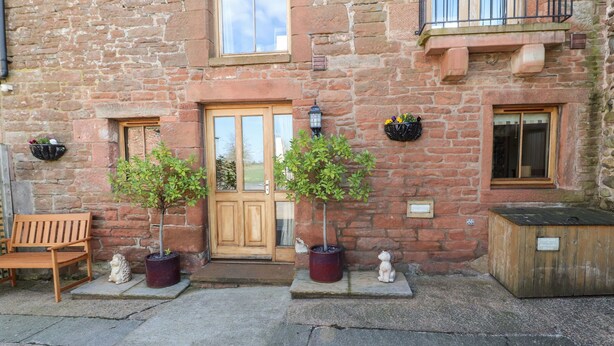 A stone building with a wooden door, two potted trees, a bench, and a small statue. - Swallow's Nest (Penrith)