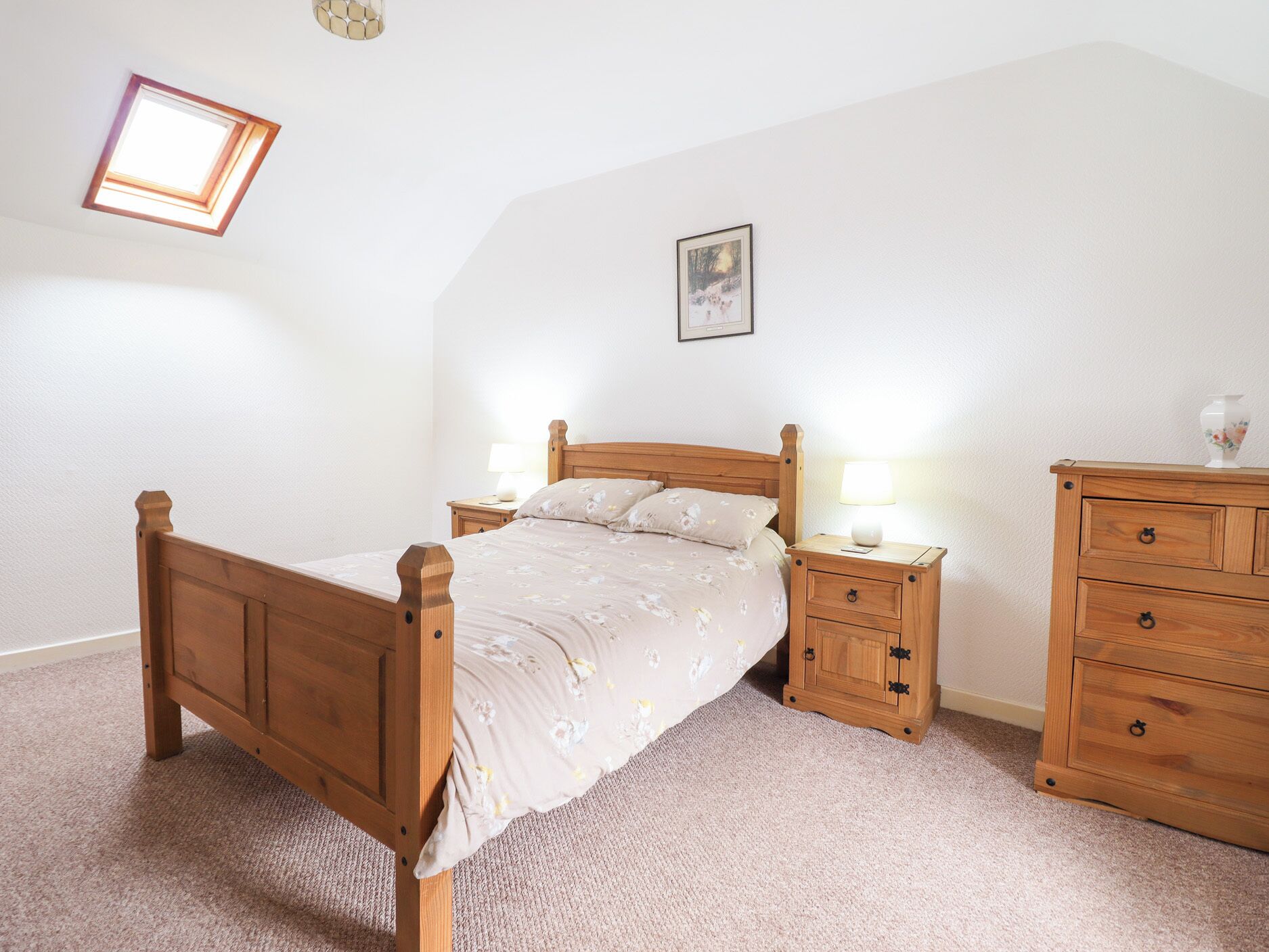 Une chambre à coucher avec un lit en bois, deux tables de chevet avec des lampes, une commode, un tableau encadré au mur et une lucarne.