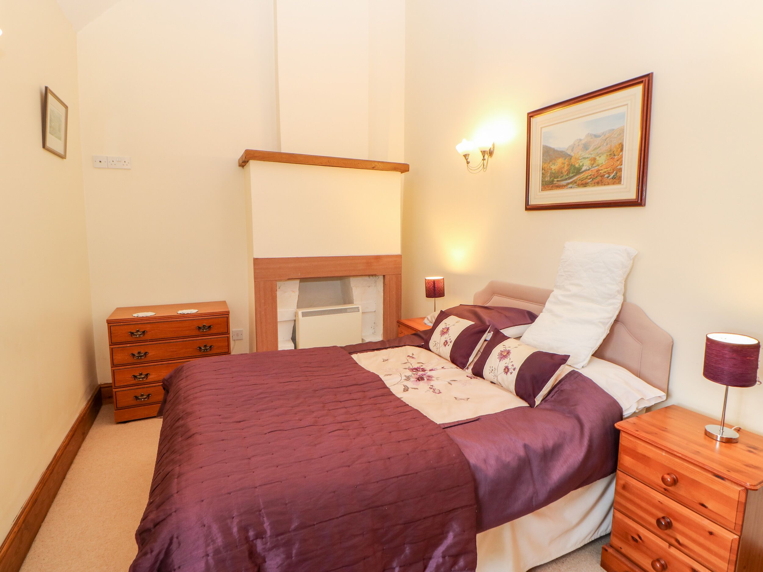 Een slaapkamer met een bed, een kaptafel, een nachtkastje, een wandlamp, een ingelijste foto en een open haard.