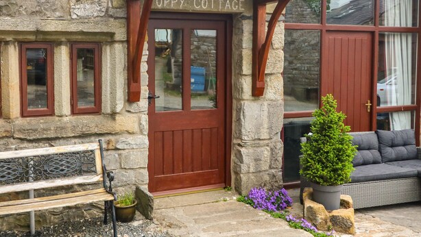 Une petite maison en pierre avec une porte rouge portant l’inscription « OPPY COTTAGE », un banc, une plante en pot et un canapé gris.