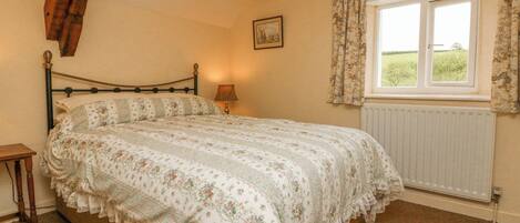 A bedroom with a bed, a nightstand, a lamp, a window with curtains, a radiator, and a picture on the wall.
