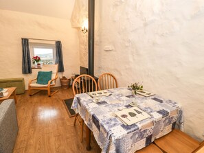 A cozy dining area with a table set for two, wooden chairs, a green cushion, a wicker basket, and a view through a window with curtains.