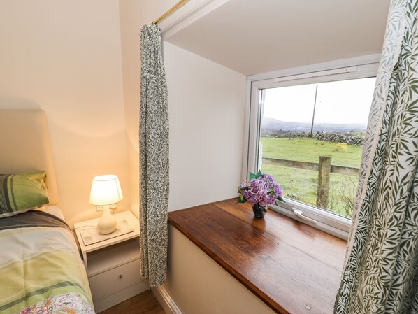 A bedroom with a bed, a bedside table with a lamp, a window with a view of a grassy field, and a curtain with a leaf pattern.