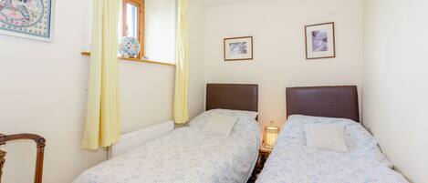Un dormitorio con dos camas individuales, una silla de madera, una ventana con cortinas y dos cuadros en la pared.