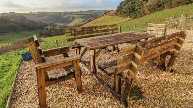Une table et des bancs en bois rustique, installés sur un sol de gravier, offrent une vue pittoresque sur des collines verdoyantes et un ciel dégagé.