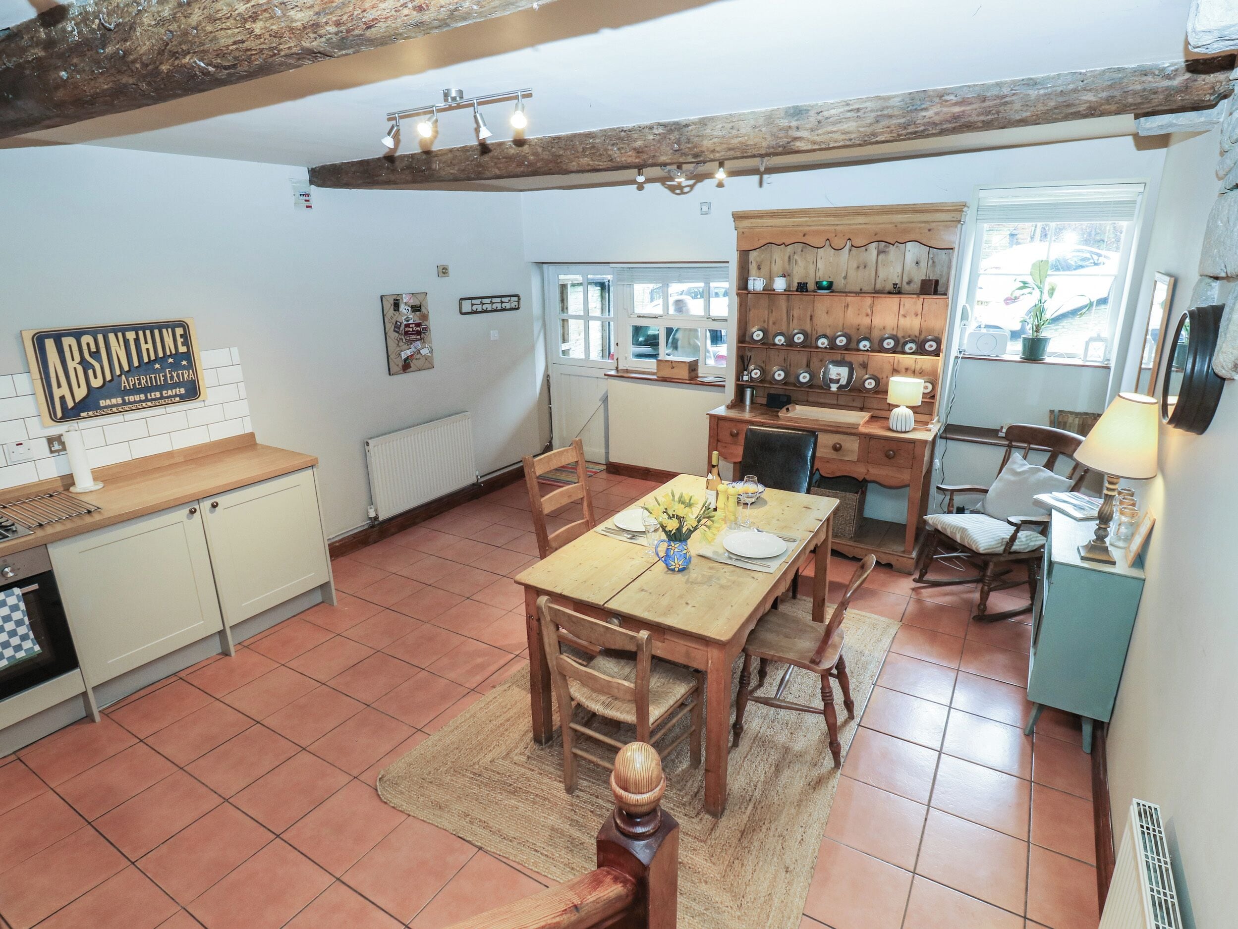 A rustic kitchen with a wooden table set for dining, a tiled floor, and a stone wall with a sign.