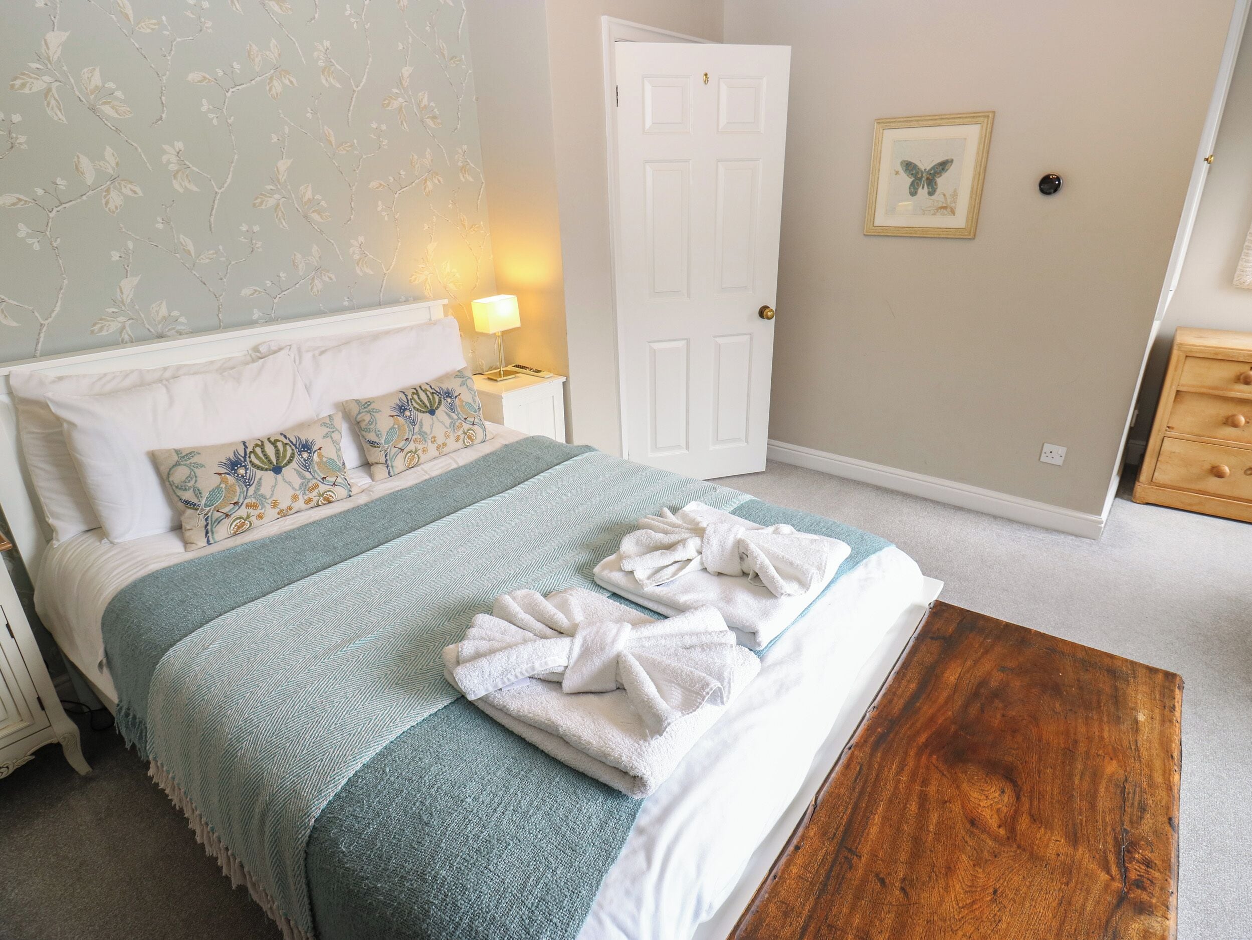 Una cama bien hecha con una colcha azul y blanca, una mesita de noche de madera con una lámpara, una puerta blanca y un pequeño aparador de madera.
