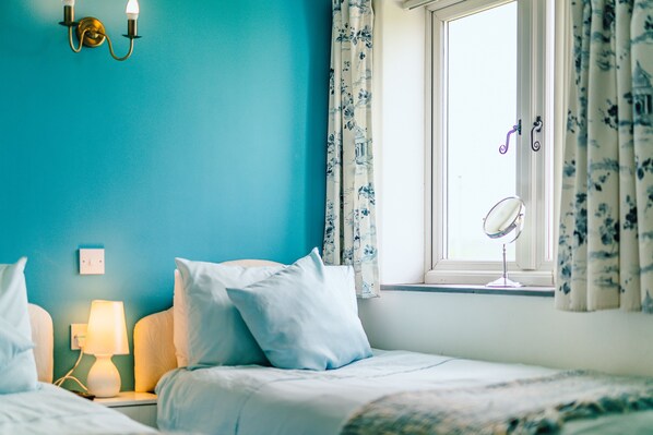 A bedroom with a teal wall, a bed with white bedding, a bedside lamp, a window with floral curtains, and a mirror on the windowsill. - Crown Cottage (Holywell)