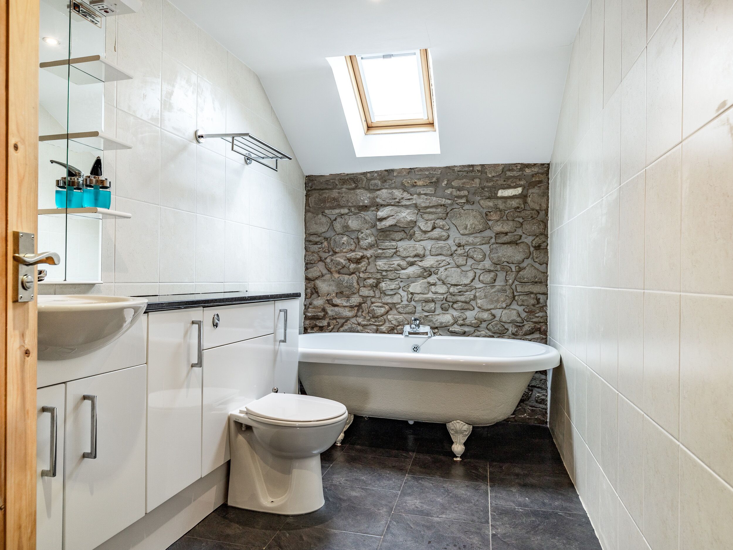 Un baño compacto con pared de piedra, una bañera independiente, inodoro y lavamanos con espejo.