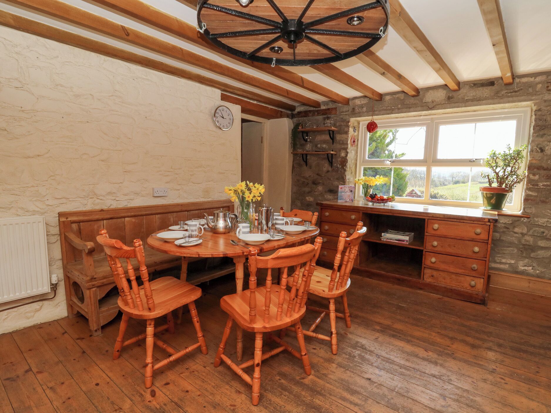 A dining area with a wooden table set for four, surrounded by wooden chairs. A rustic ceiling fan is visible, and a window with a view of greenery is present.