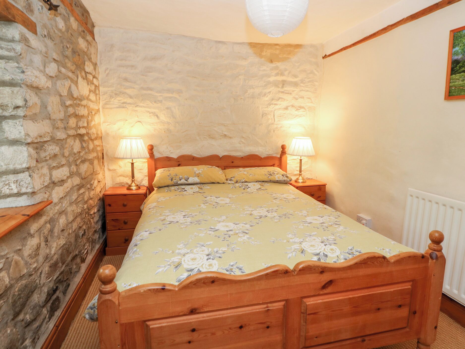 A bedroom with a wooden bed, two bedside tables with lamps, a stone wall, and a radiator.