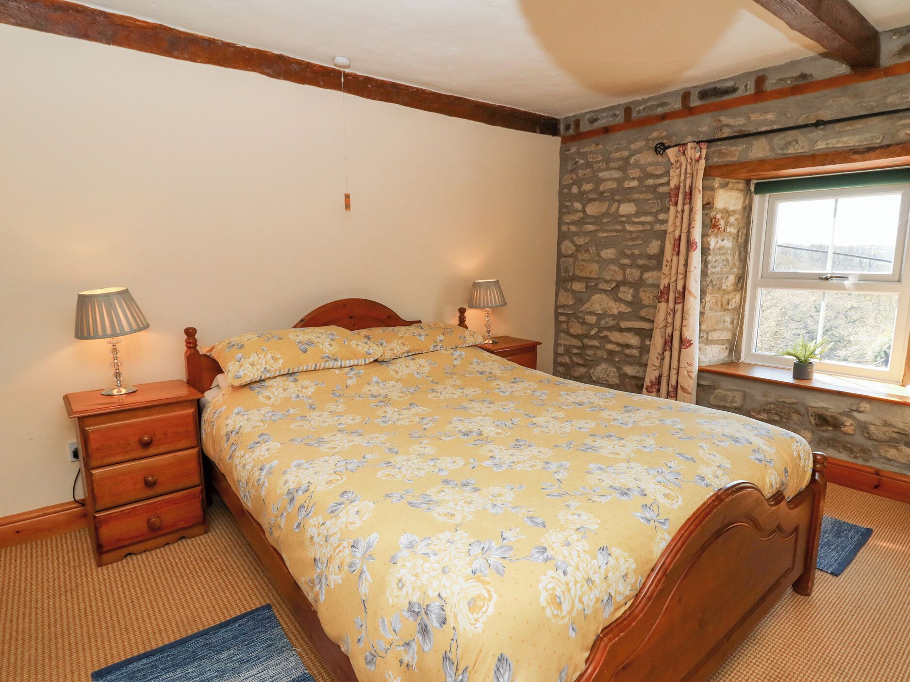 A bedroom with a wooden bed, two bedside tables with lamps, a stone wall, and a window with curtains.