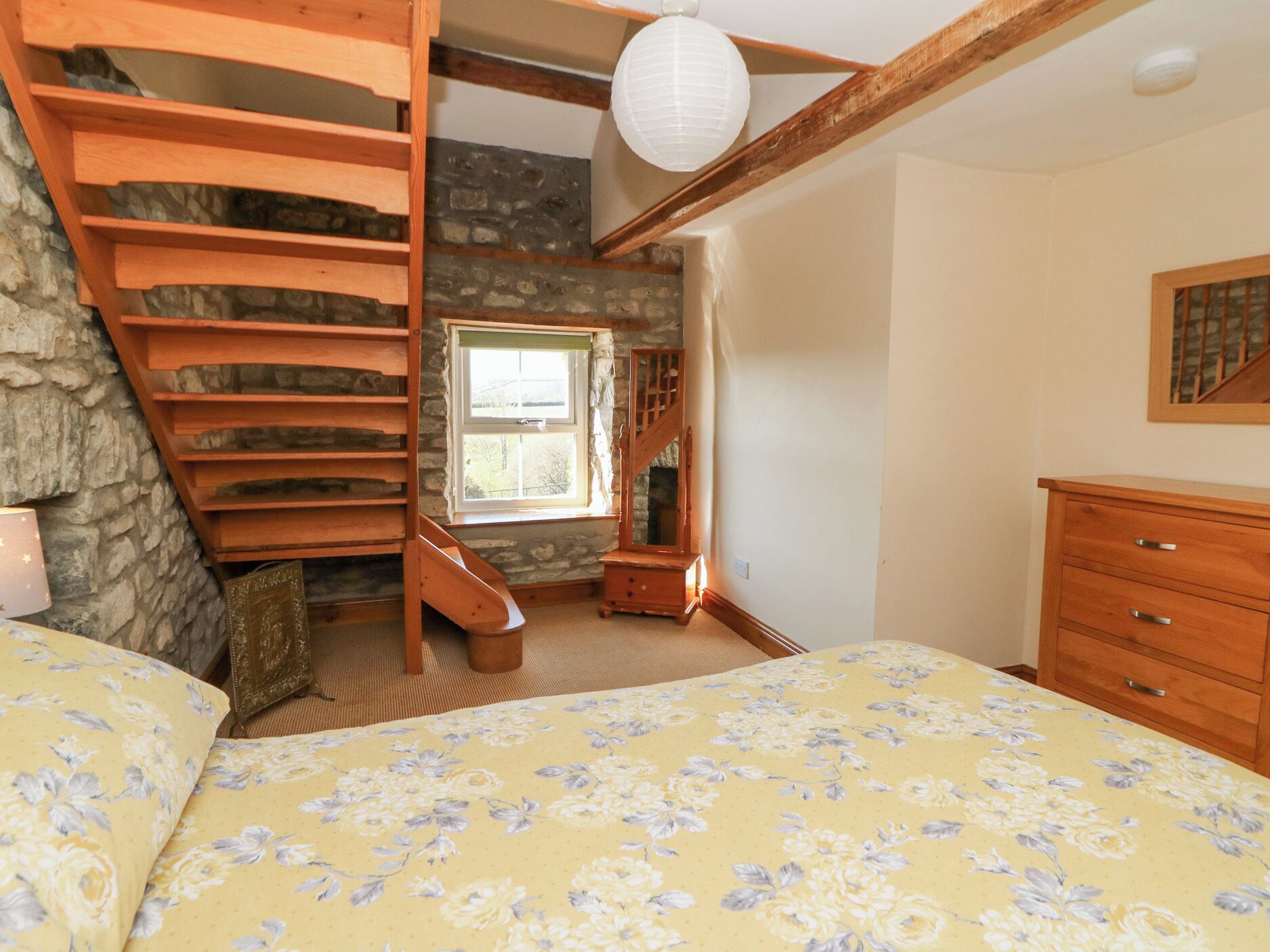 Une chambre avec un escalier en bois, un lit recouvert d’une literie à motifs floraux, une commode et un mur en pierre.