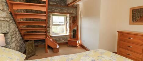 Une chambre avec un escalier en bois, un lit recouvert d’une literie à motifs floraux, une commode et un mur en pierre.
