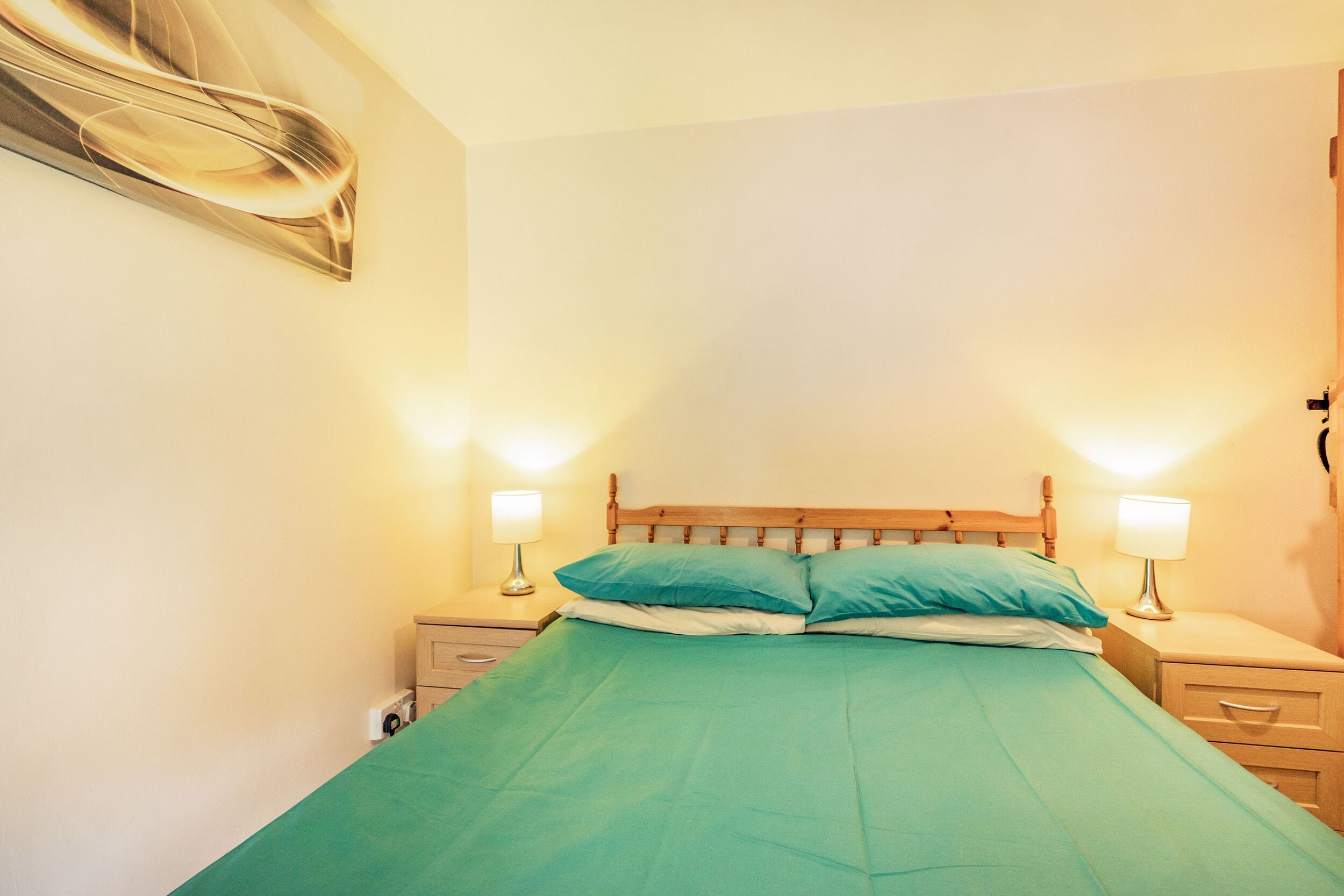 Een net opgemaakt bed met een groene sprei en twee kussens, met aan weerszijden nachtkastjes met lampen, in een kamer met effen muren en een houten hoofdeinde.