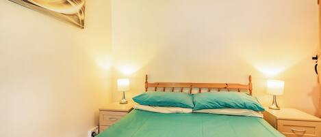 Een net opgemaakt bed met een groene sprei en twee kussens, met aan weerszijden nachtkastjes met lampen, in een kamer met effen muren en een houten hoofdeinde.