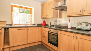 A modern kitchen with wooden cabinets, a stainless steel oven, and a window with a view of greenery. - The Granary (Thirsk)