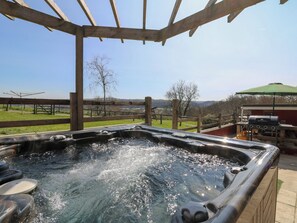 A hot tub with a wooden pergola overhead, surrounded by a grassy area with a view of hills and a picnic table.