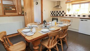 A dining area with a wooden table and chairs, a kitchen with a checkered backsplash, and a window with a view of the outdoors. - Mill Cottage (Lampeter)