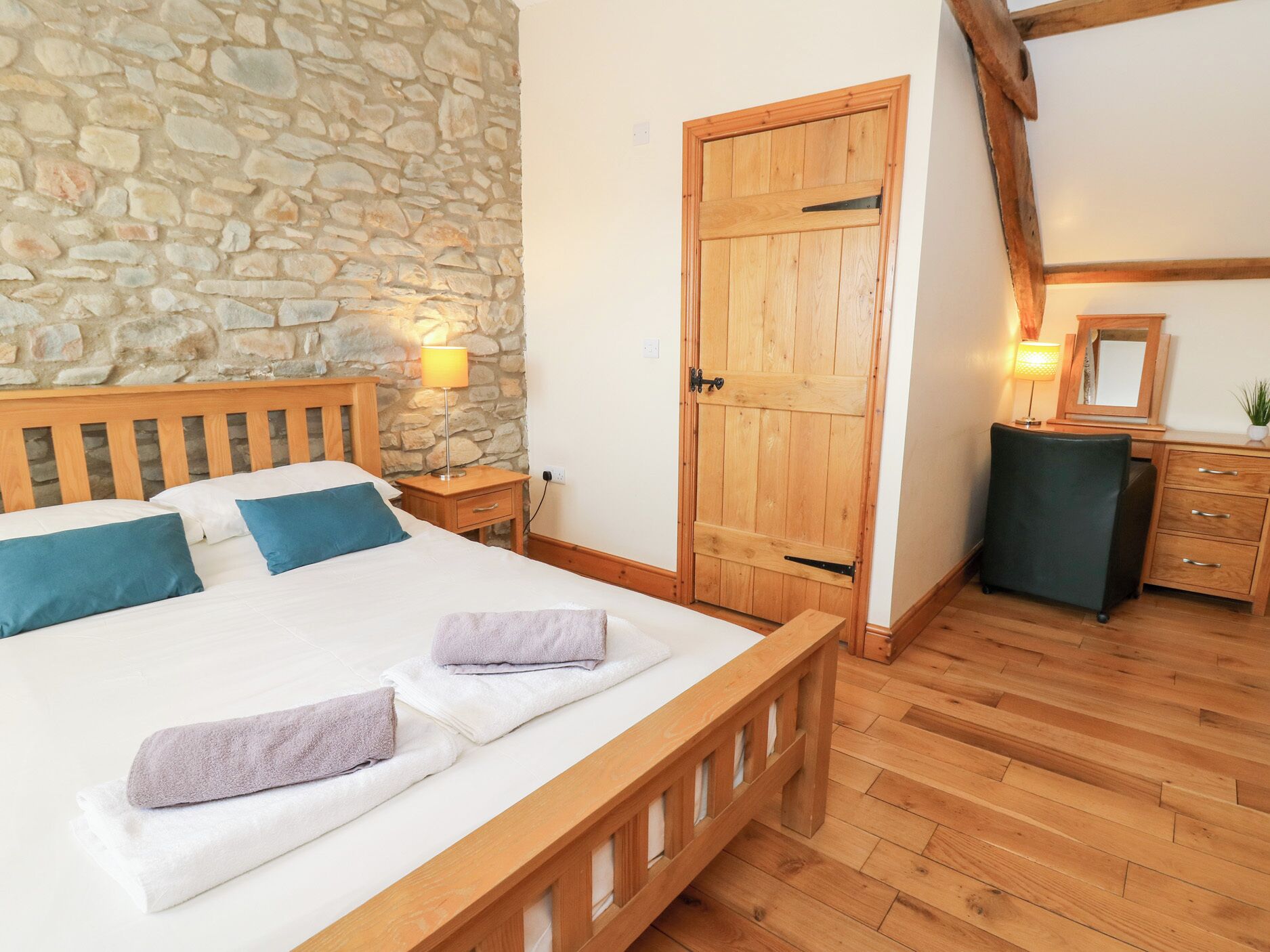 A bedroom with a stone wall, wooden bed, bedside tables, a wooden wardrobe, and a desk with a mirror.