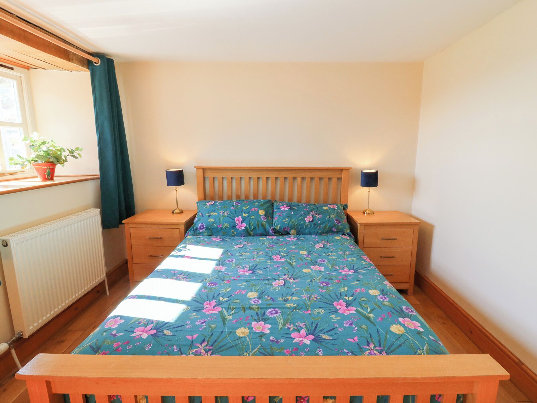 Un dormitorio con cama de madera, ropa de cama floral, dos mesitas de noche con lámparas, una ventana con cortinas y un radiador.