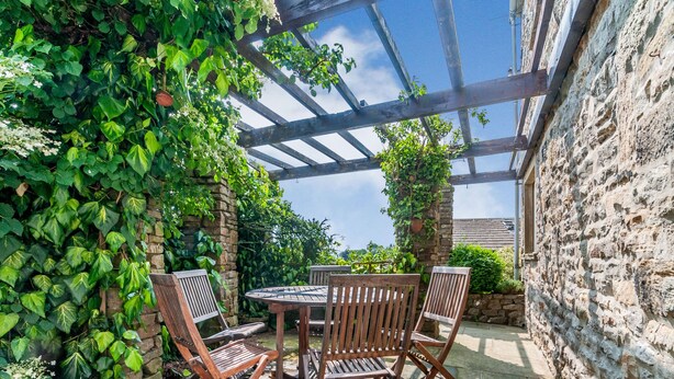 An outdoor patio with a wooden table and chairs, surrounded by stone walls and lush greenery. - The Shippon (Skipton)