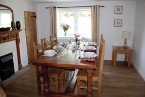 A dining room with a wooden table set for six, flanked by wooden chairs, a fireplace, and a window with curtains. - Manor Farm Cottage (Swaffham)