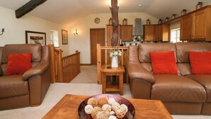 A living room with two brown leather sofas, a wooden coffee table with a decorative bowl, wooden beams on the ceiling, and a kitchen in the background. - Copper Beech Cottage (Coniston)