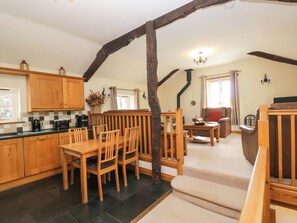 A cozy kitchen with wooden cabinets, a dining table, and chairs. There is a staircase leading to a living area with a sofa and a coffee table.