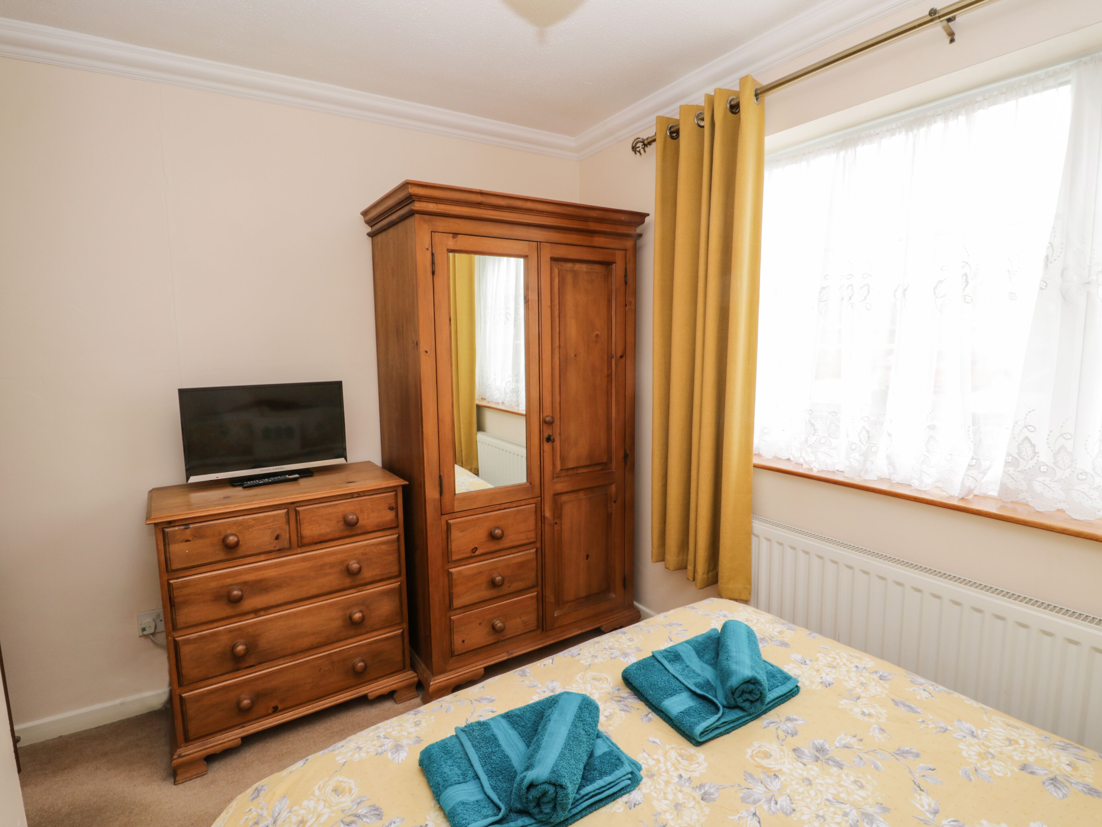 Une chambre à coucher avec un lit, une armoire en bois, un téléviseur posé sur une commode et une fenêtre avec des rideaux.