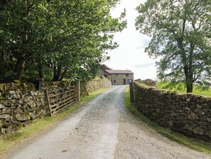 A rural road leading to a stone house with a wooden gate, surrounded by stone walls and trees.