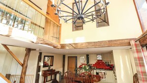 A spacious dining area with a high ceiling and a chandelier, wooden beams, and a view of the upper floor through a glass railing. - The Barn (Thetford)