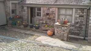 A stone building with a tiled roof, a small porch with potted plants, and a window with shutters. - Courtyard Cottage (Kendal)