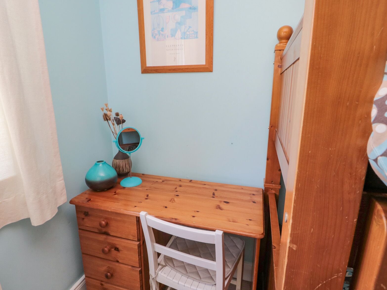 Quarto pequeno e bem arrumado com cômoda de madeira, uma cadeira branca, uma mesinha e um quadro na parede.