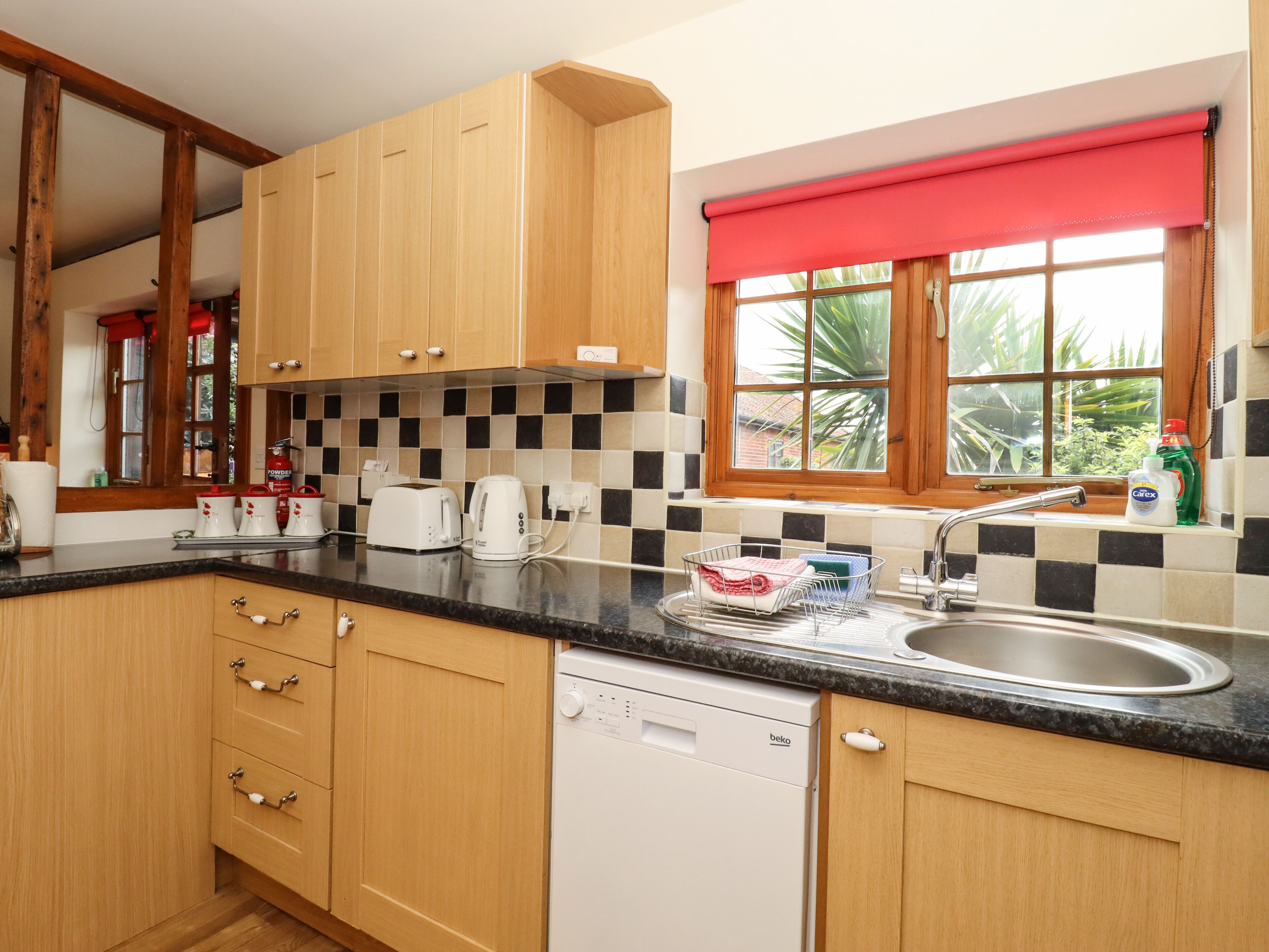 A kitchen with wooden cabinets, a black countertop, a dishwasher, and a window with a red blind.