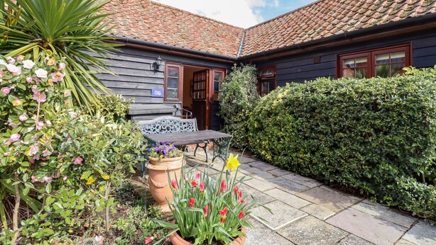 A courtyard with a bench, potted plants, and a house with a tiled roof. - Poppy Cottage (Woodbridge)