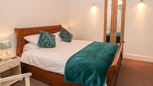 Cama bem arrumada com cabeceira de madeira, duas almofadas decorativas, manta azul-turquesa, mesa de cabeceira com abajur e espelho de corpo inteiro.