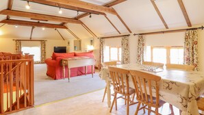 A spacious room with a high ceiling and exposed wooden beams, a dining area with a table and chairs, a sofa, and a television. - Hayloft Cottage (Woodbridge)