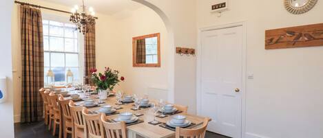 A dining room with a long table set for six, wooden chairs, a chandelier, and a window with curtains.