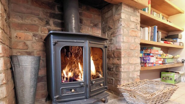 A wood-burning stove with visible flames, a metal bucket, and a wicker basket on a stone floor. - The Lodge (Scarborough)