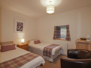 A bedroom with two beds, a window with curtains, a radiator, a desk with a lamp, and a chair.