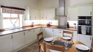 A modern kitchen with white cabinets, a wooden dining table set for four, and a built-in oven. - Beudy (Corwen)