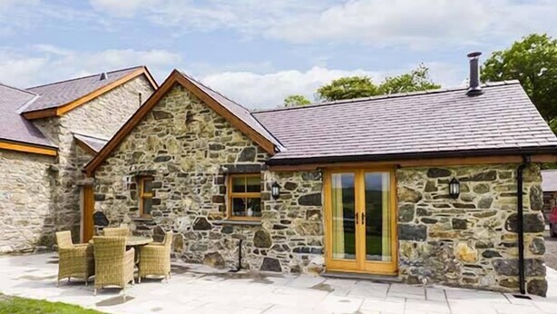 A stone building with a sloped roof, a wooden door, and a small patio with wicker furniture. - Beudy (Corwen)