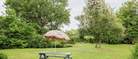 A picnic table with a thatched umbrella in a lush garden with dense trees and a well-maintained lawn.