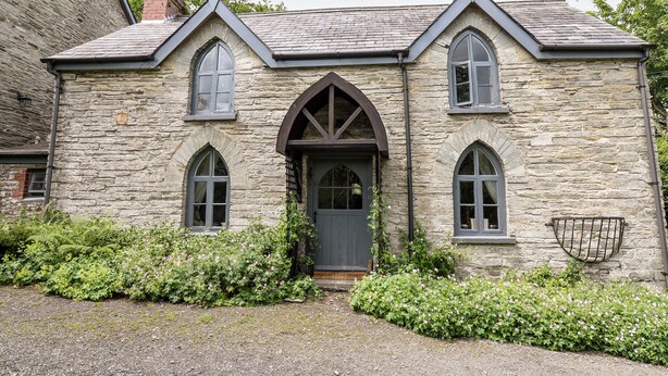 A stone building with a dark entrance, arched windows, and a small garden area with pink flowers. - Maesgwyn (Cardigan)