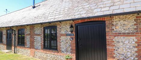 A brick building with a black door, a sign reading "Keepers Cottage," and a small garden area with a potted plant and a metal grate.