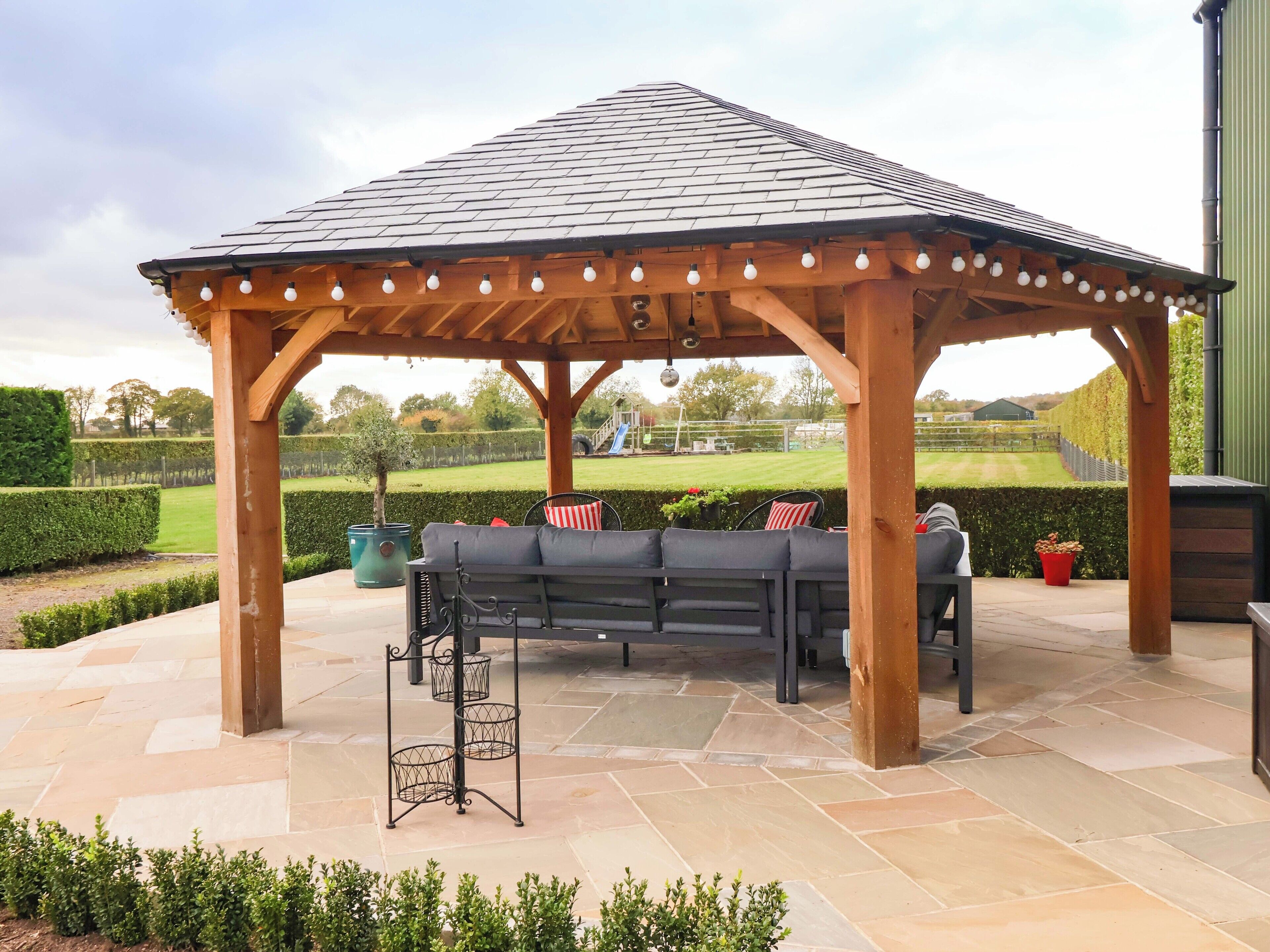 A wooden gazebo with a tiled roof and string lights, a black outdoor sofa, and a metal table with baskets.