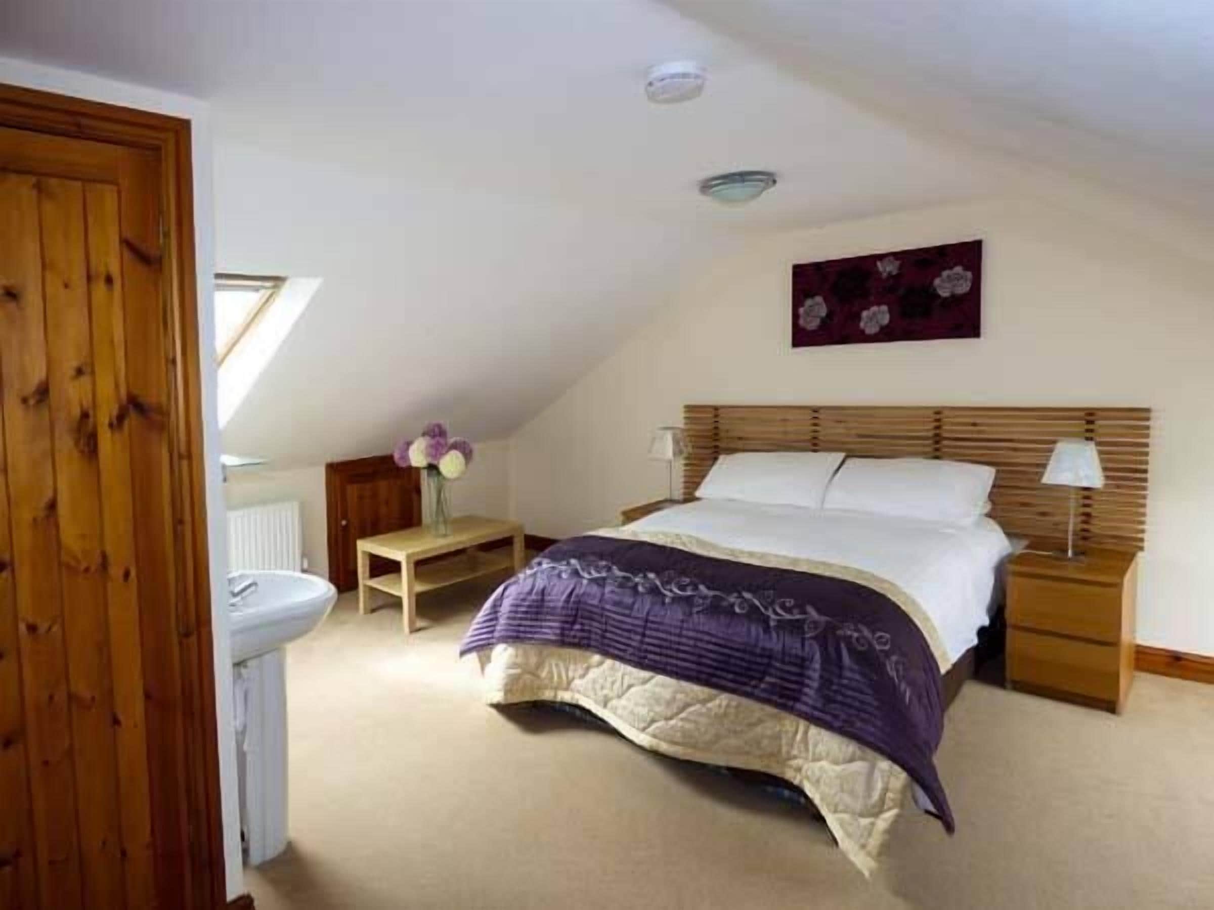 Een slaapkamer met een schuine kap, een houten bed, een nachtkastje met een lamp, en een klein nachtkastje met een bloemenvaas.
